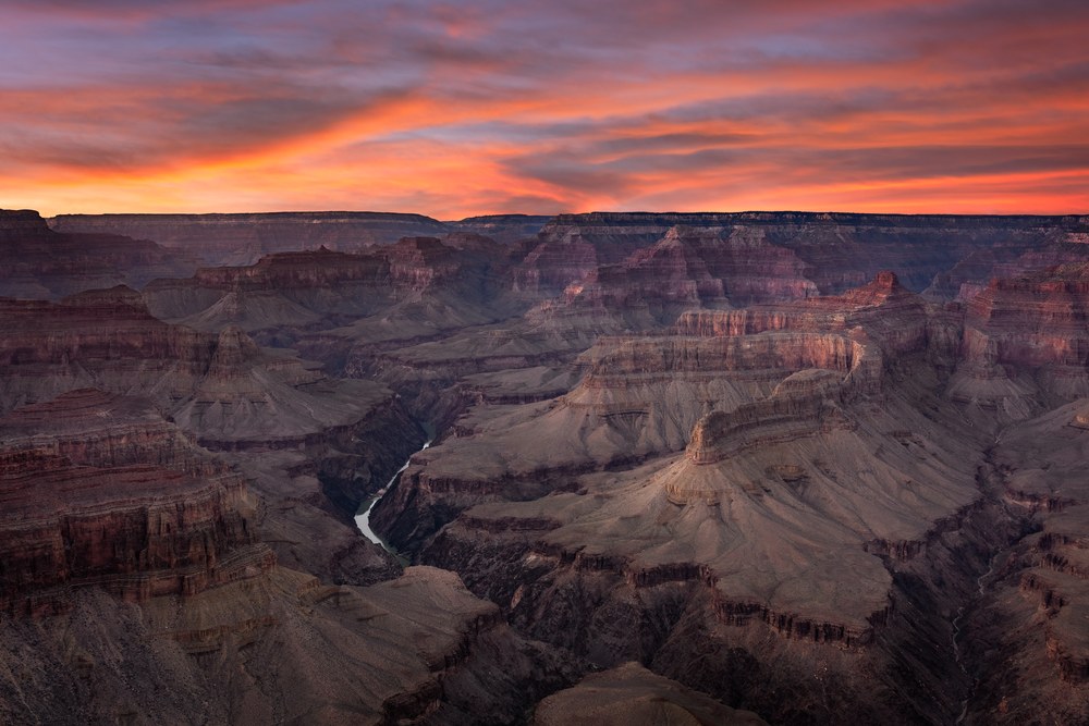 Burning orange cloud sits over a wide canyon, a thin stretch of river visible in the shadowed gorge below.
