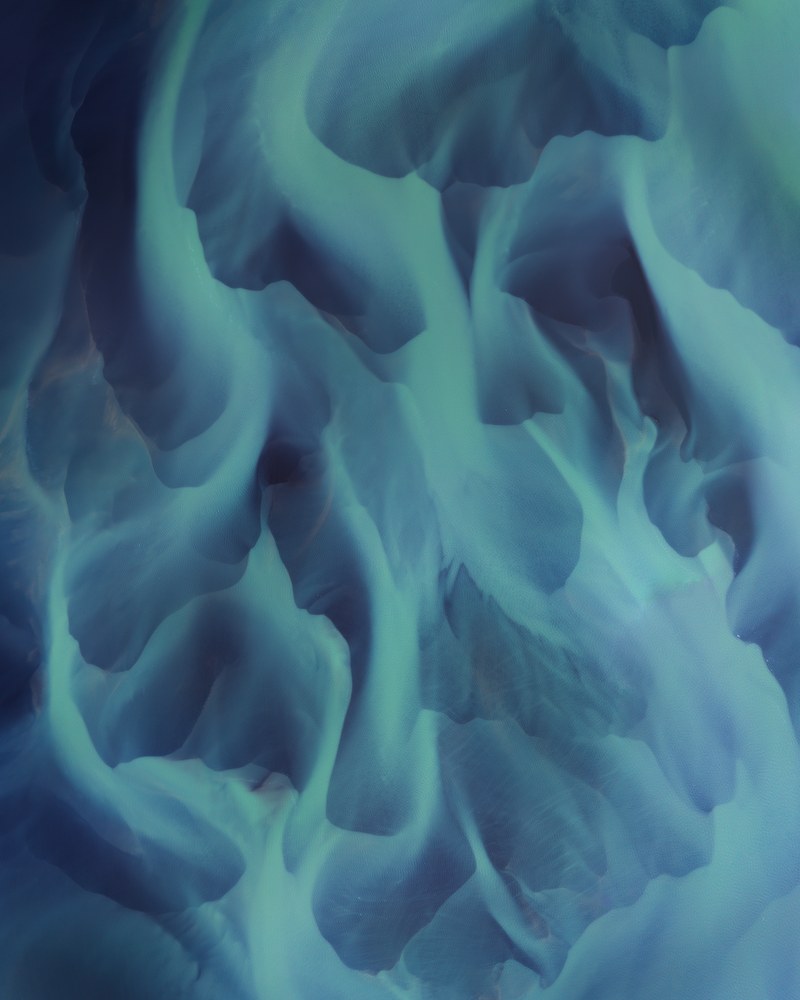 An aerial abstract of a braided glacial river, pale green channels branching like flames across blue-grey silt.