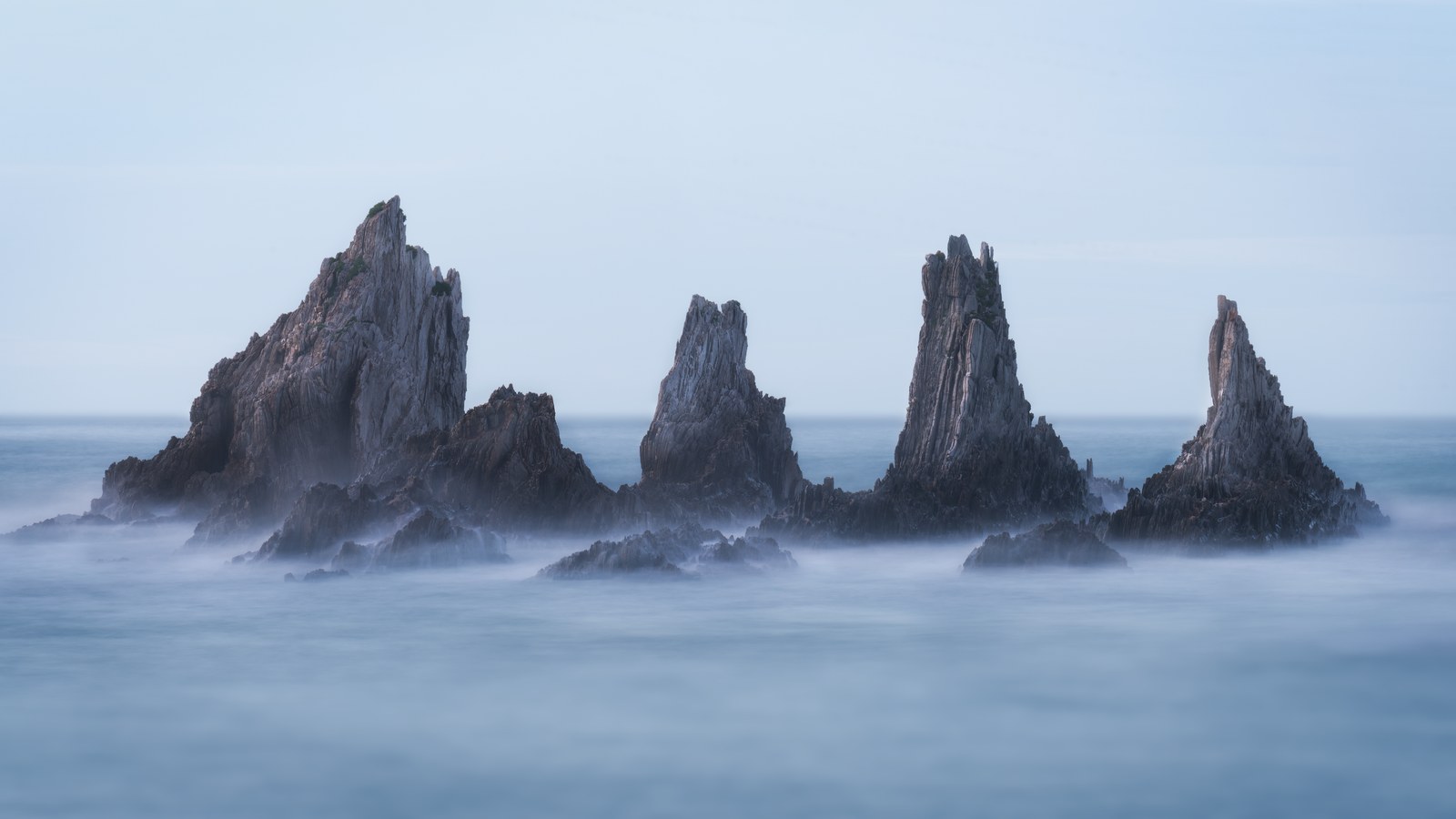 Four sharp rock spires stand in a pale, mist-blurred sea, their bases dissolved by the long exposure.