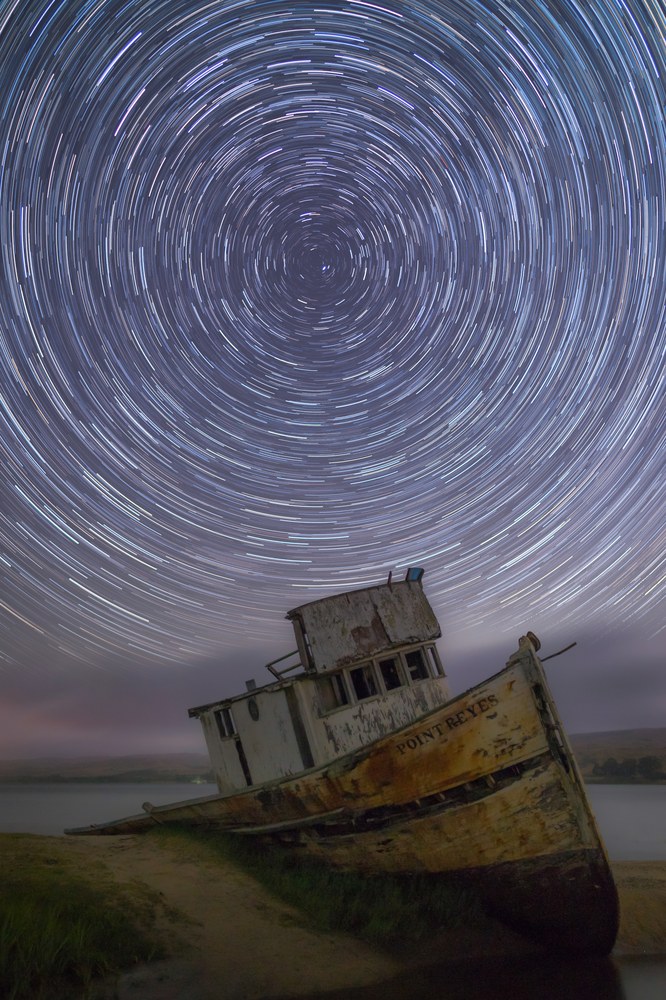Concentric star trails wheel overhead above a beached wooden fishing boat, its hull weathered and listing in the grass.