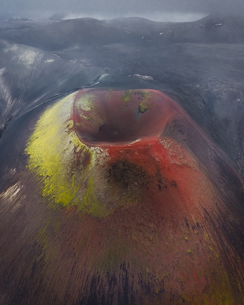 An aerial of a cinder cone whose crater rim is stained deep red and sulphur yellow against surrounding black ash.