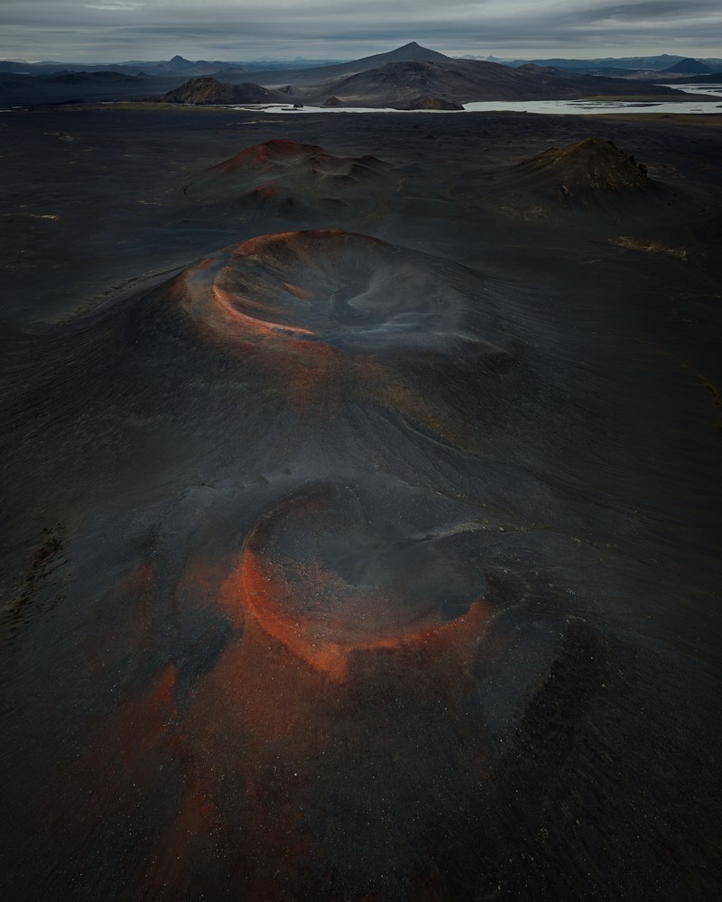Two rust-red cinder cones rise from a black ash field, their rims catching low light, hills fading into cloud beyond.
