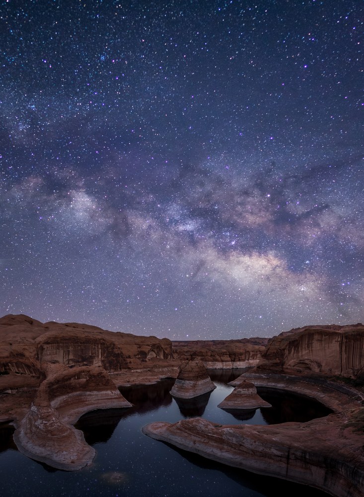 The Milky Way arcs over a flooded canyon where pale sandstone fins stand mirrored in still, dark water.
