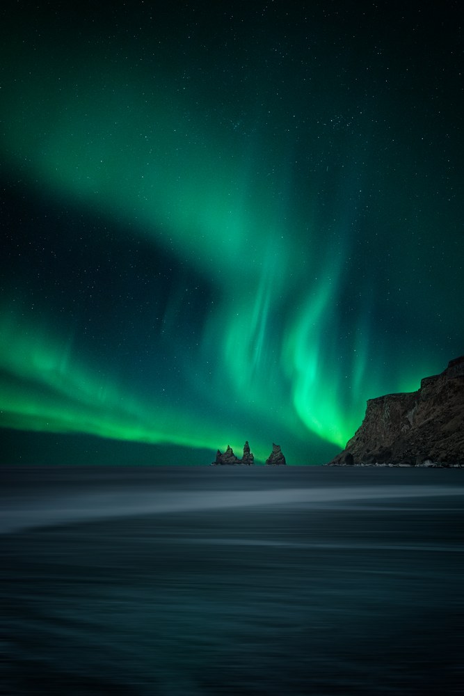 Green aurora sweeps across a star-filled sky above a dark headland and distant sea stacks, the ocean below smoothed to a sheet.