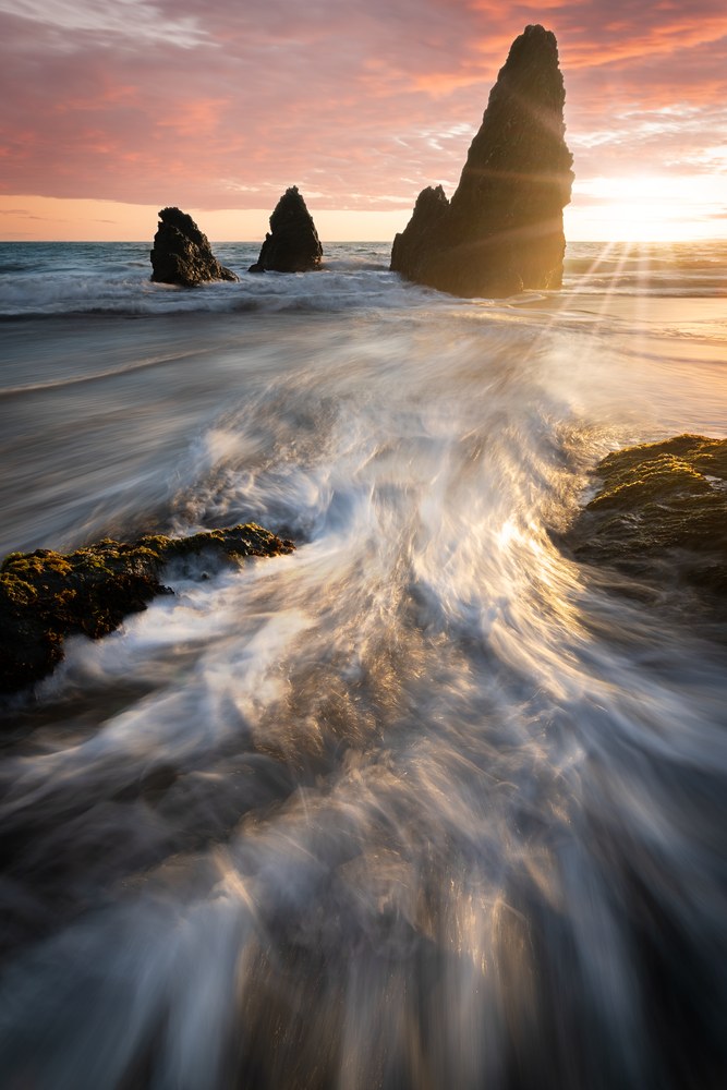Backwash streams toward the camera across mossy rock while a low sun flares between three dark sea stacks under pink cloud.