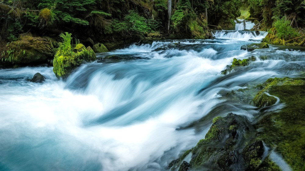 A pale blue river runs hard over mossy rock, a smaller waterfall visible upstream through the trees.