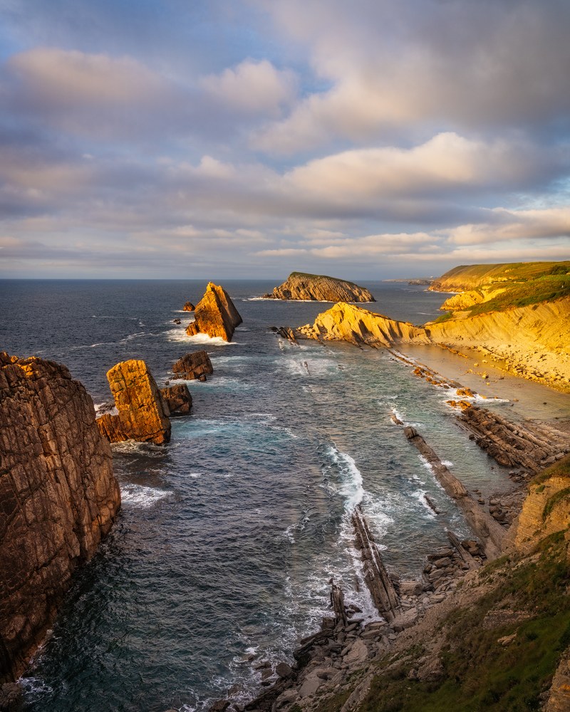 From a clifftop, low sun lights tilted slabs of rock stepping down into a cove where sea stacks stand in choppy water.