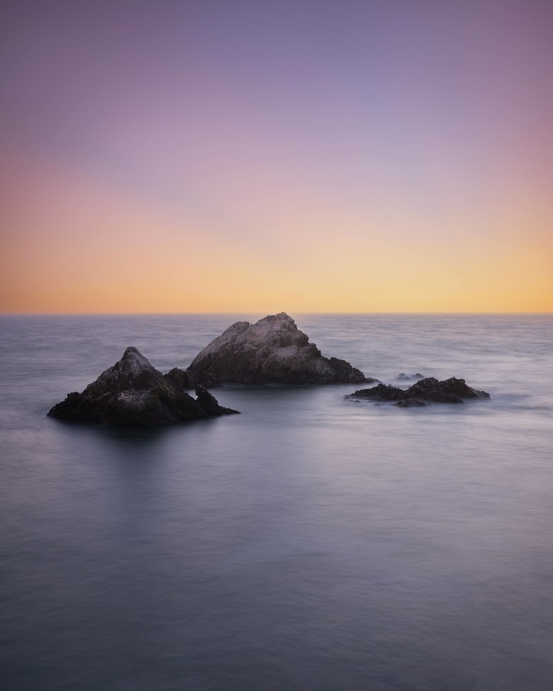 Two low rock clusters sit in a glassy long-exposure sea beneath a gradient sky running from lavender down to amber.