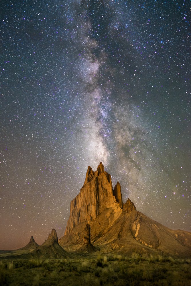 A jagged volcanic plug rises from flat scrub, warm-lit, with the Milky Way standing almost vertically above it.