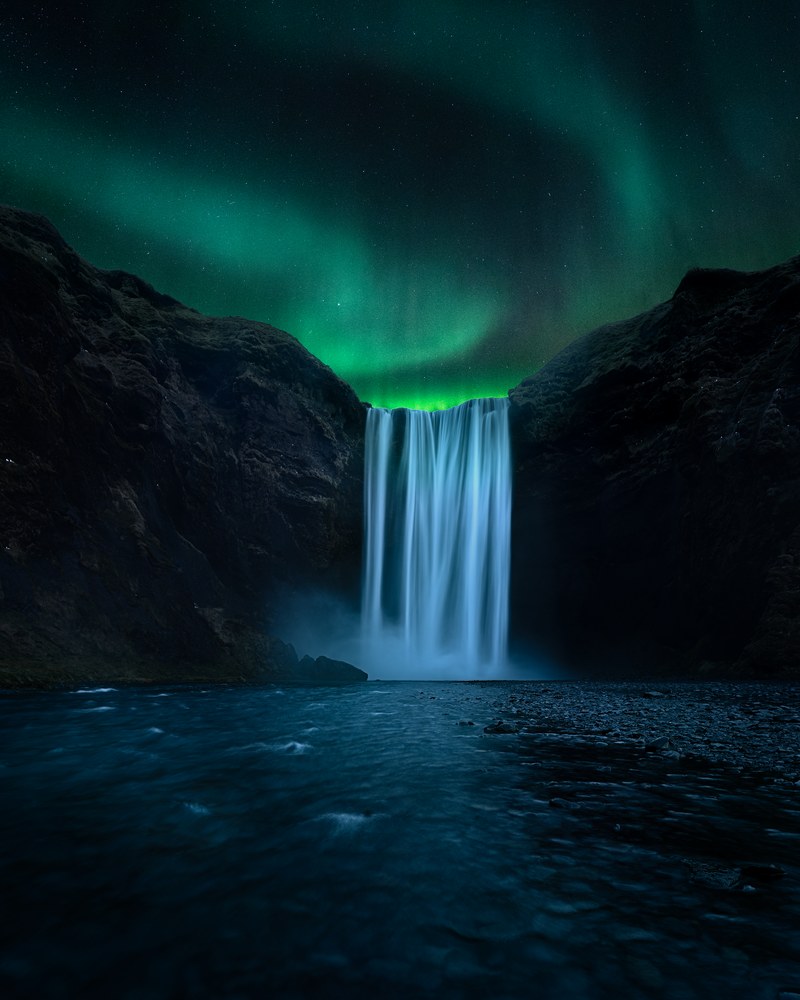 A broad waterfall glows between dark cliffs at night as green aurora arcs overhead, the river running black in the foreground.
