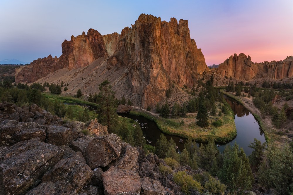 A river bends around a tan rock massif with a jagged crest, dark boulders in the foreground and pink sky beyond.