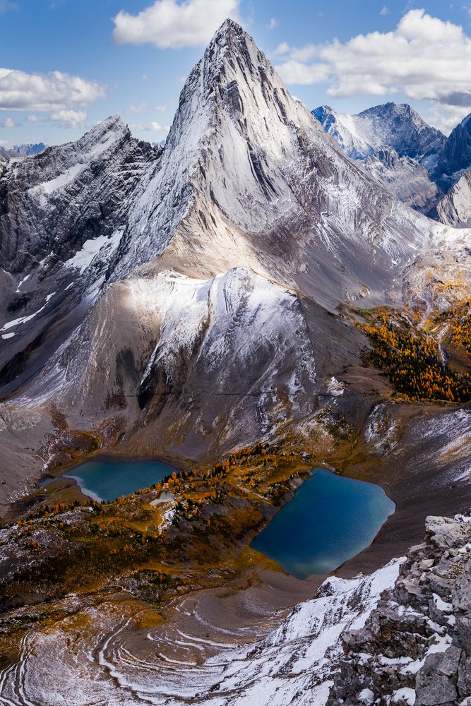 From high on a ridge, a sharp pyramid peak stands above two teal tarns ringed with golden larches and fresh snow.