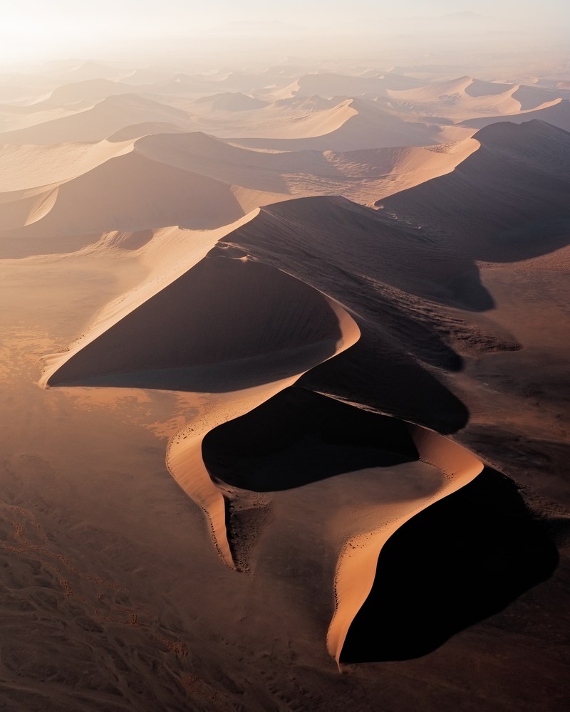 An aerial of linked dunes whose lit crests curve away in a chain, their leeward faces dropping into black shadow.