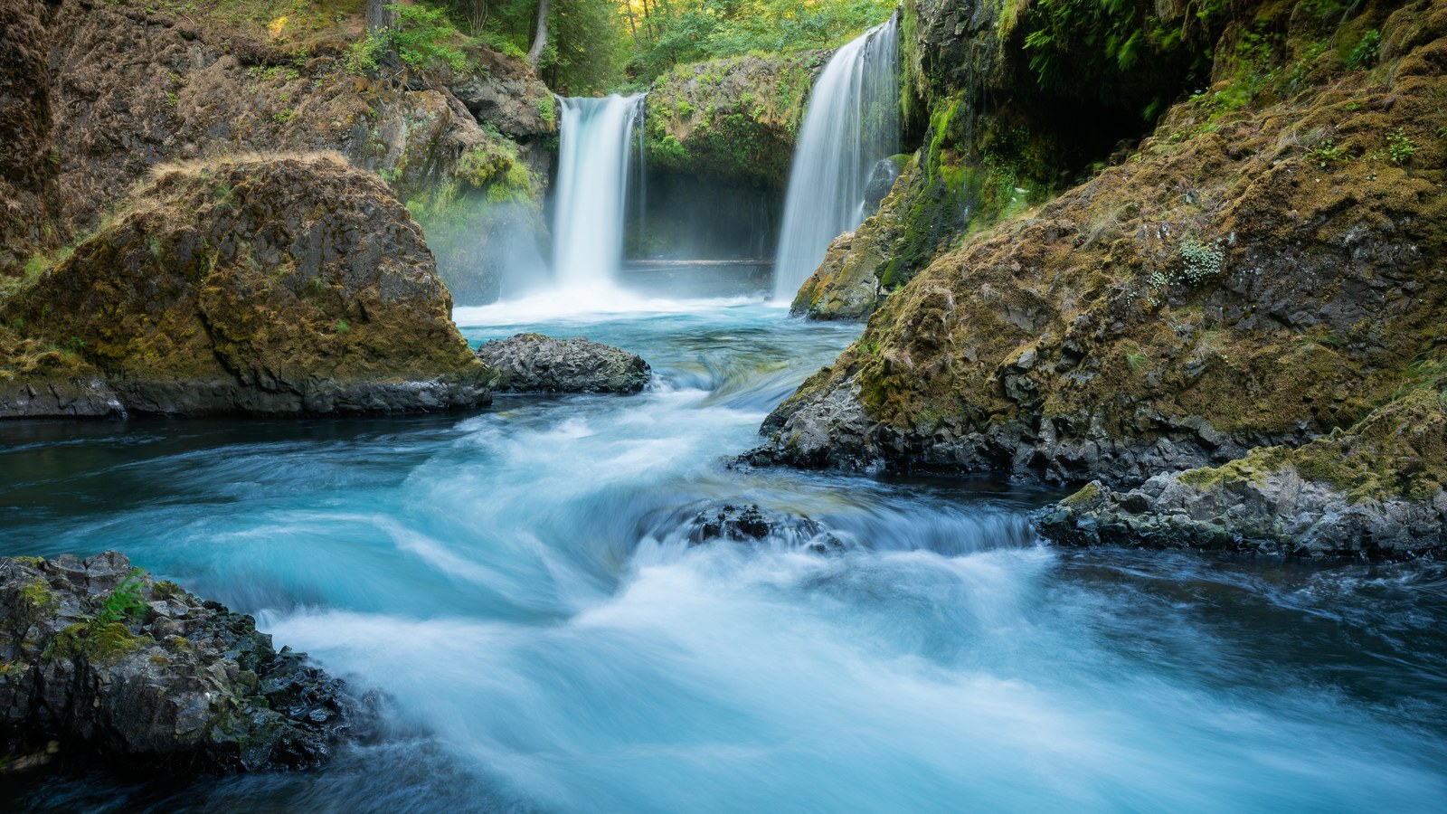 Two waterfalls drop between mossy basalt walls into a turquoise river that rushes over rock toward the camera.