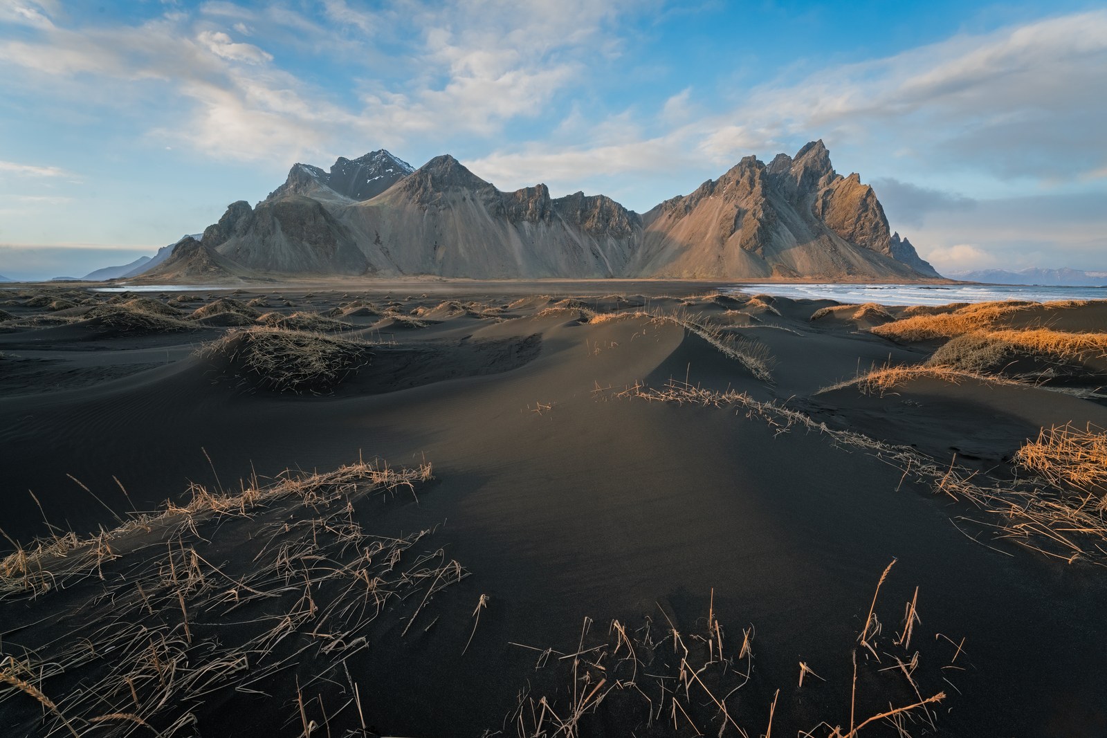 Black sand dunes tufted with dry grass run toward a jagged dark mountain massif under a blue sky streaked with cloud.