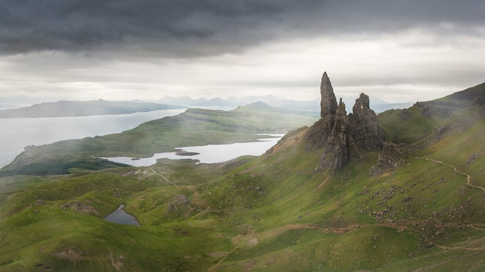 Rock pinnacles stand on a green ridge above a sea loch, distant hills fading under a heavy grey cloud ceiling.