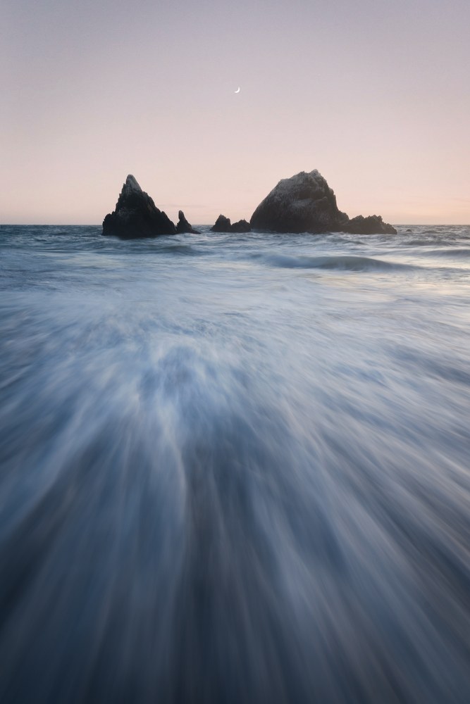 Water rushes seaward in radial streaks toward two rock clusters, a thin crescent moon hanging in the pink dusk sky above.