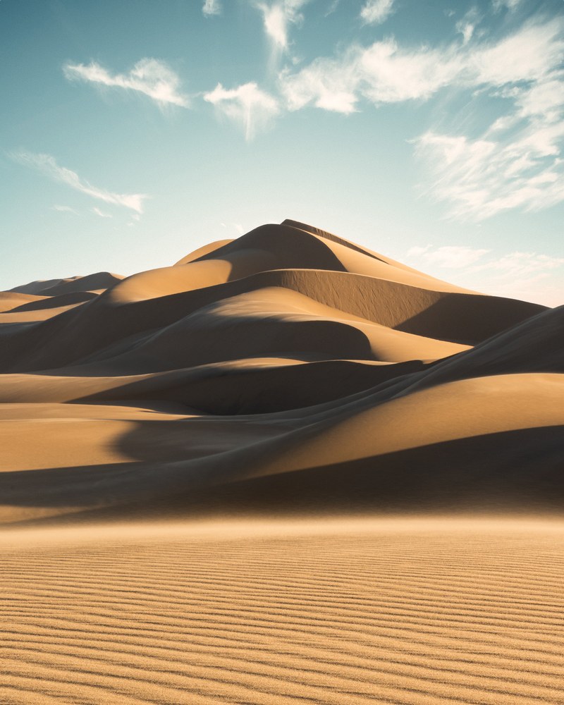 Pale dunes stack into ridges and sharp shadow lines under a blue sky, wind ripples running across the flat sand in front.