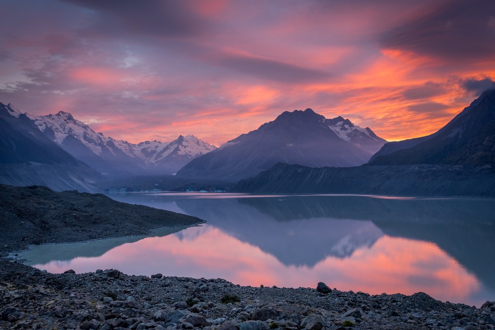 Pink sunrise cloud burns above snow-capped peaks and repeats in a still glacial lake, moraine rubble in the foreground.