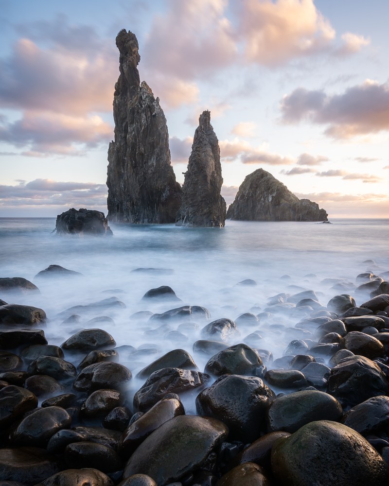 Two slender sea stacks and a flatter third rise past a foreground of large rounded boulders, the water between them blurred to mist.