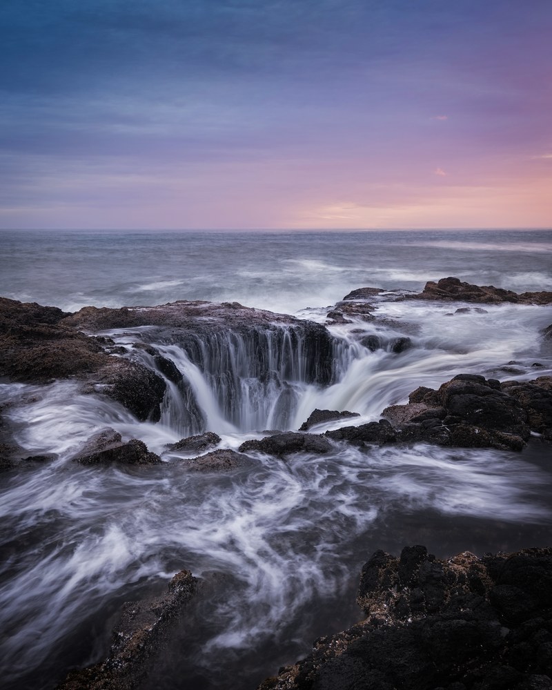 Seawater pours over the lip of a hole in a basalt shelf while surf swirls across the surrounding rock under a pastel dusk sky.