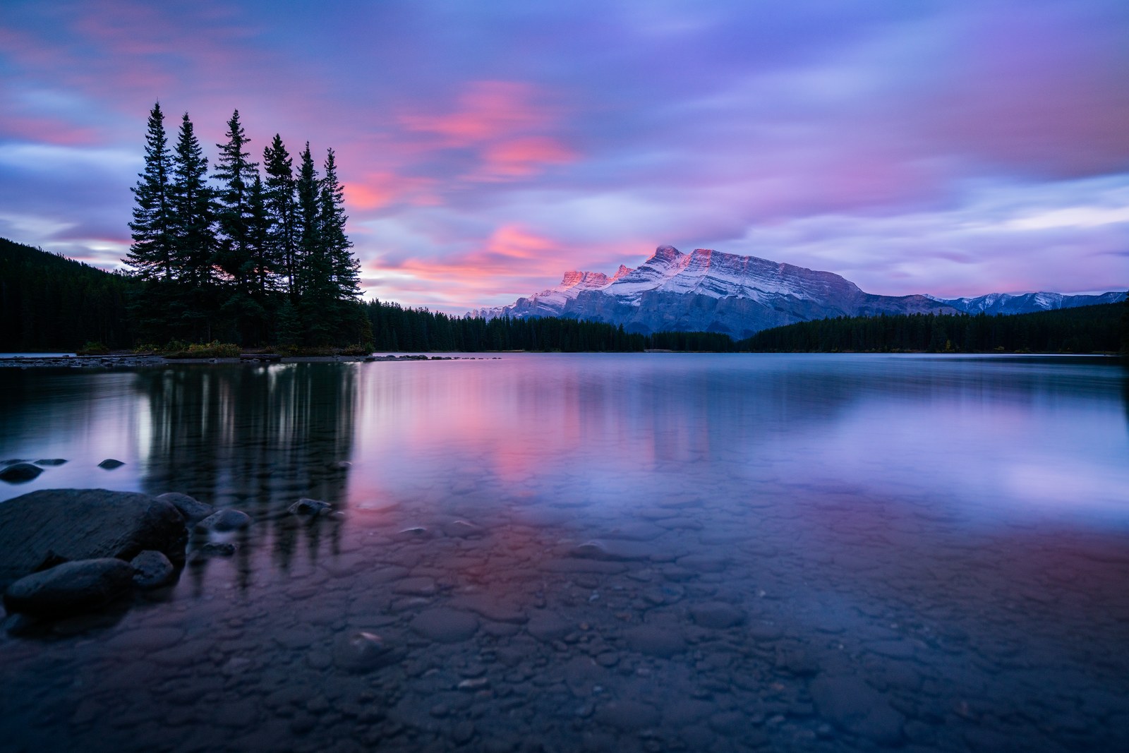 Pink and purple dusk cloud sits over a broad mountain and a stand of spruce, stones visible through the still lake in front.