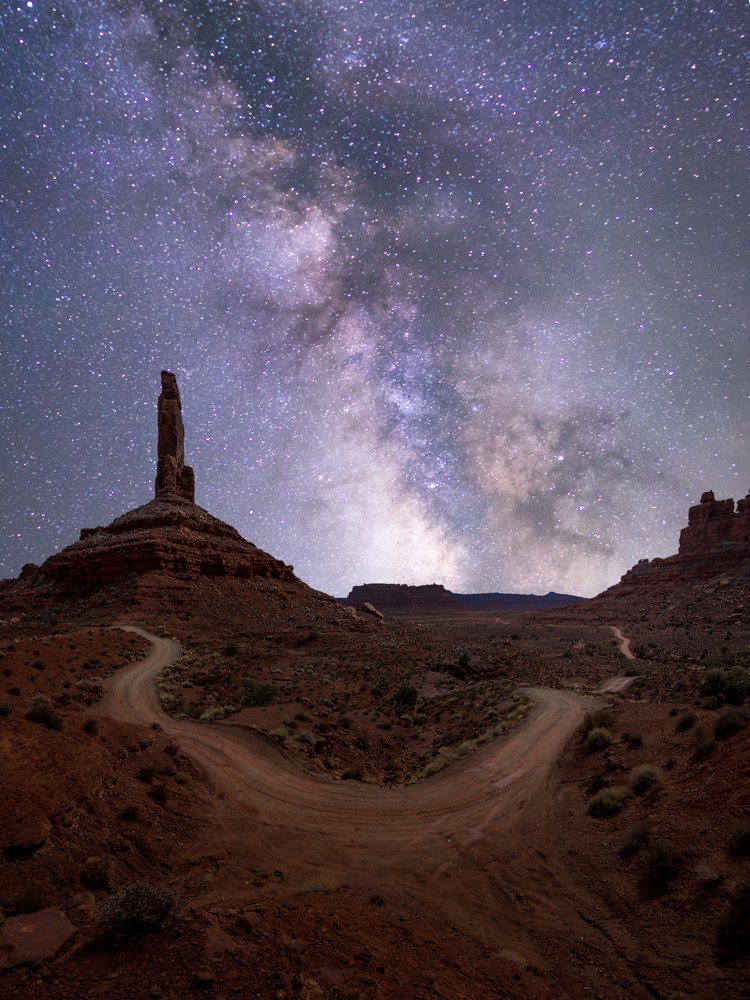 A dirt road forks in the foreground below a red butte topped by a slender spire, the Milky Way filling the sky.
