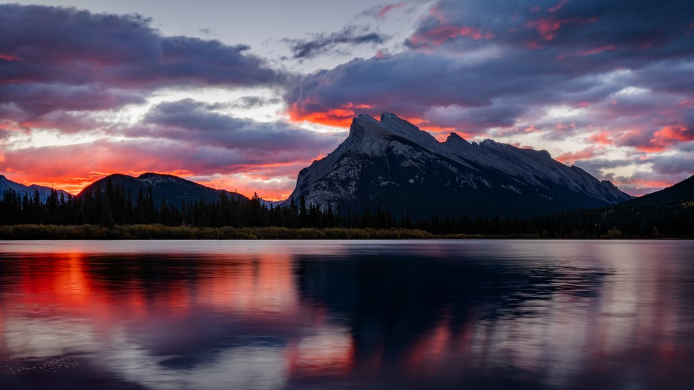 Fiery red cloud burns over a broad mountain and repeats in the still lake below it.
