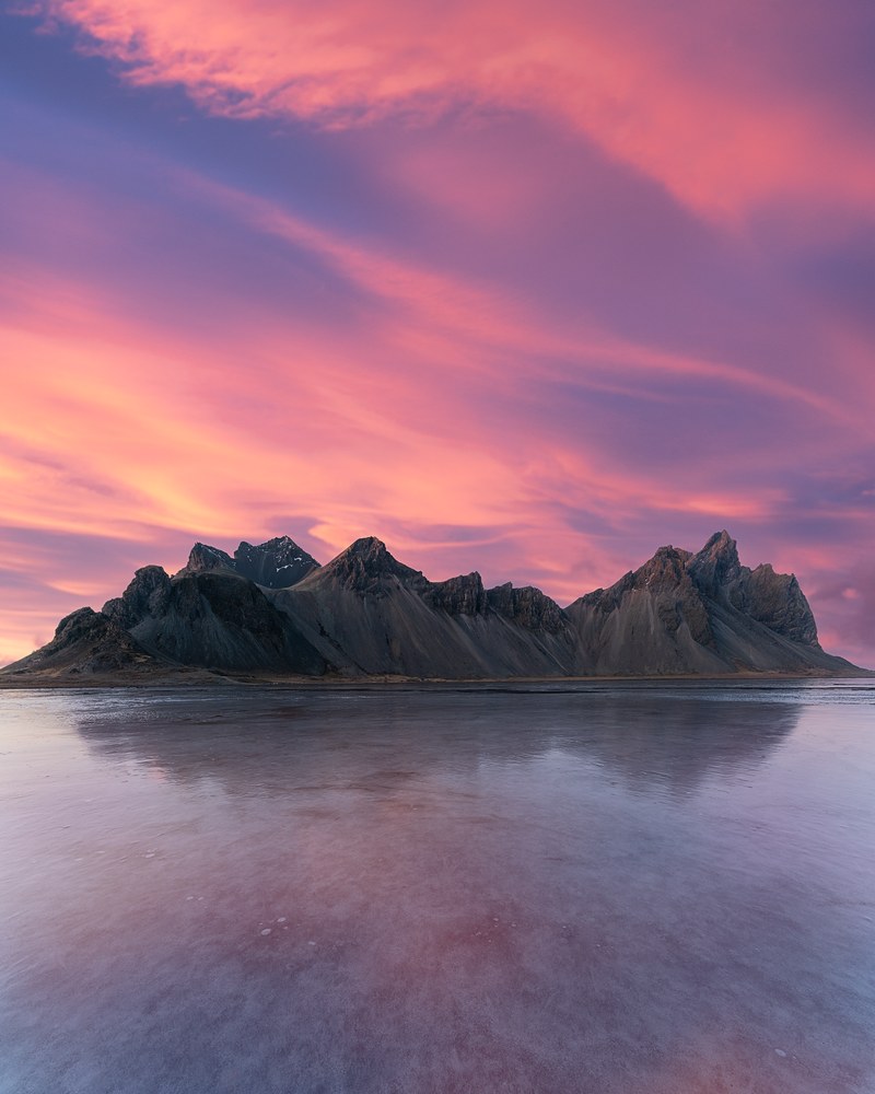 A jagged dark mountain massif sits low on the horizon beneath a sky of pink and magenta streaked cloud, mirrored in wet sand below.
