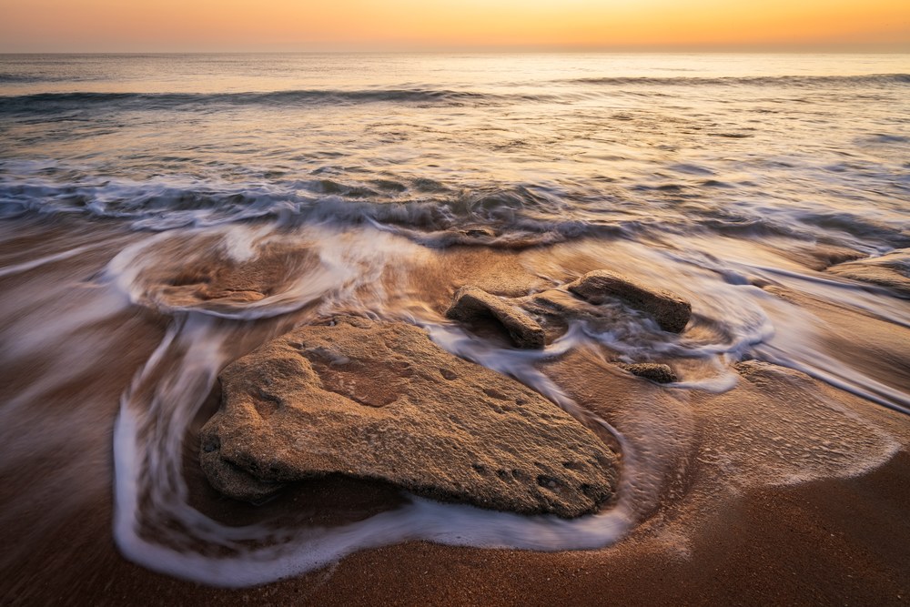 Low over a pitted coquina outcrop in shallow surf, foam curling around its edges, an orange sunrise on the horizon behind.