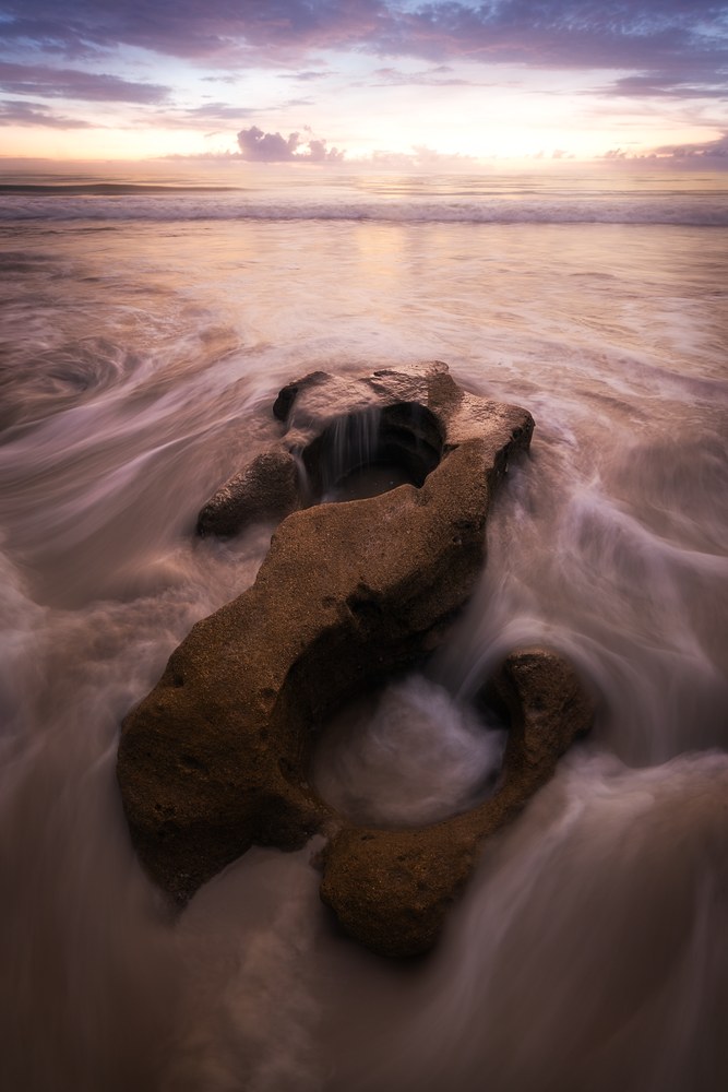 Water streams in long silky curves around a looping coquina formation on a beach, under a soft lavender and gold sunrise.
