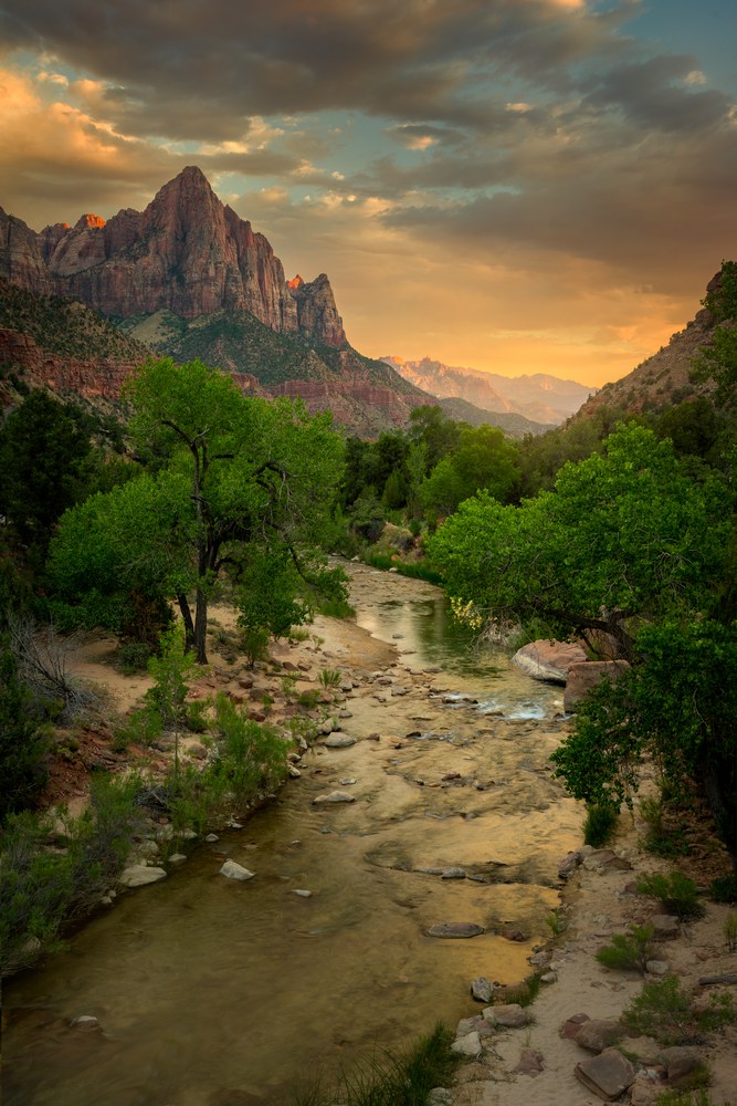 A shallow river runs between cottonwoods toward a red sandstone peak, gold cloud breaking over the ridge behind.
