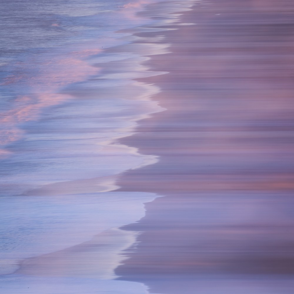 Shoreline foam blurred by camera movement into a scalloped edge between bands of lavender, pink and pale blue.