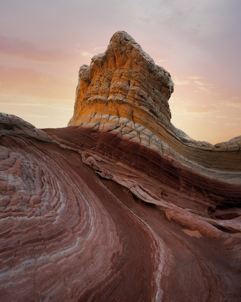 A cauliflower-textured sandstone tower rises above swirling bands of red and white slickrock under a pale pink sky.