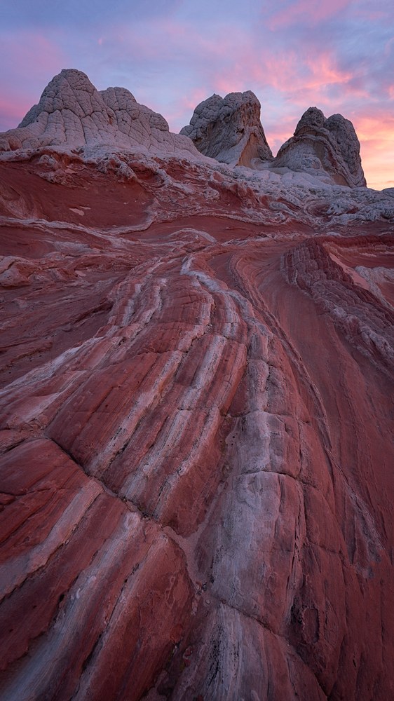 A striped ridge of red and white slickrock climbs toward pale knobbed rock towers under streaks of pink cloud.