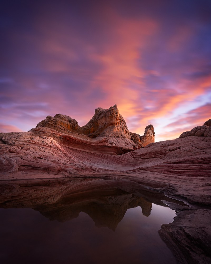 Ridged sandstone forms stand mirrored in a shallow rain pool beneath a sky of magenta and violet cloud.