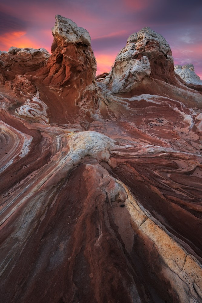 Swirled red and white sandstone ridges lead to two knobbed rock towers under a pink and violet twilight sky.