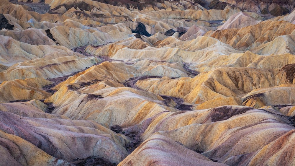 Rows of eroded badland ridges in pale yellow, pink and mauve fill the frame, dark channels running between them.