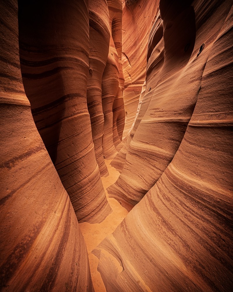 The walls of a narrow slot canyon curve overhead, banded in horizontal stripes above a smooth sand floor.